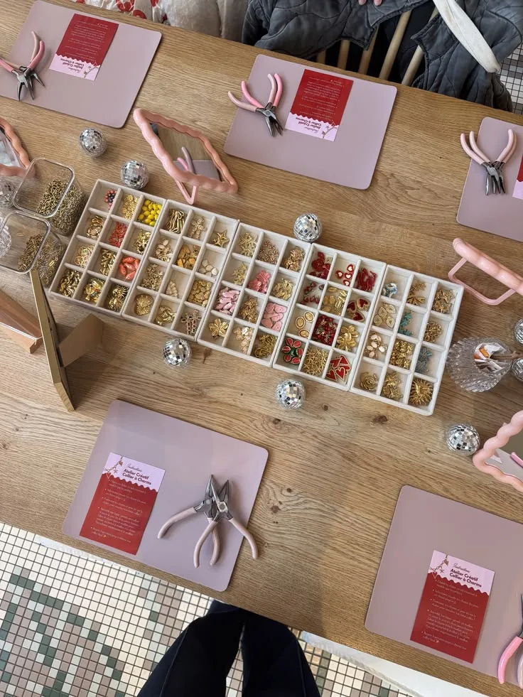 Vue du dessus d'une table d'atelier : un long plateau de charms dorés, rouges et roses, des pinces roses et de petites boules à facettes