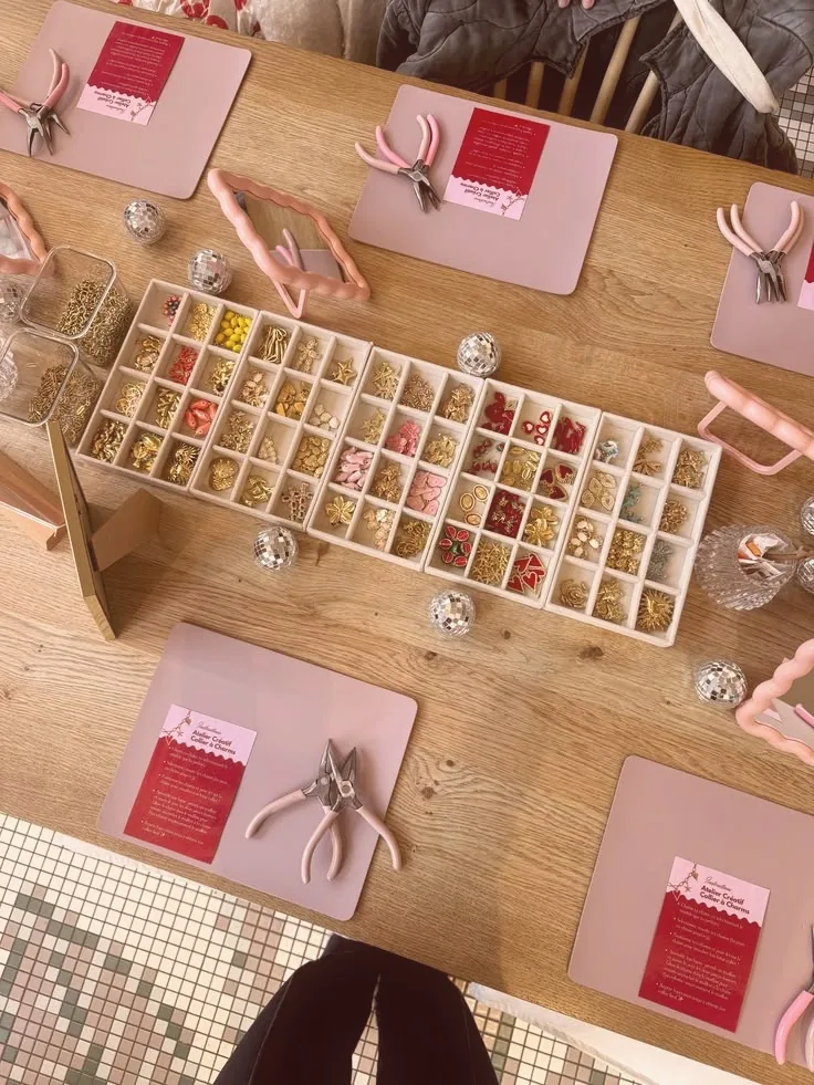 Vue du dessus d'une table d'atelier : un long plateau de charms dorés, rouges et roses, des pinces roses et de petites boules à facettes