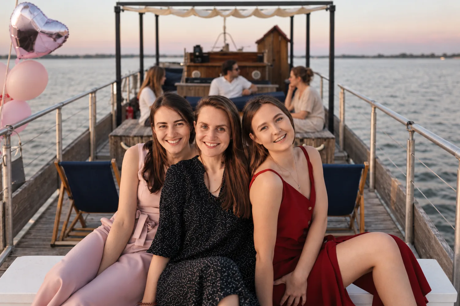 Trois amies souriantes assises à l'avant d'un bateau au coucher du soleil, des ballons roses en forme de cœur à côté
