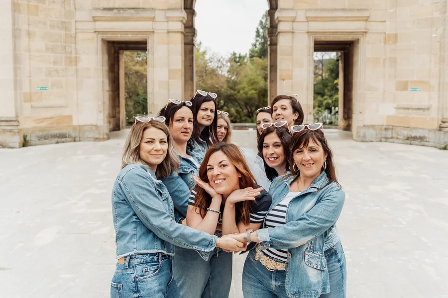 Neuf amies serrées en vestes en jean, lunettes en cœur sur la tête, l'une au centre mains sous le menton, devant un portique de pierre, EVJF à Bordeaux