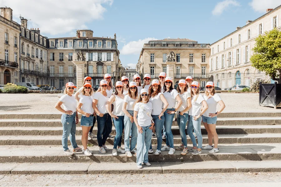 Le groupe au complet en casquettes orange, aligné sur les larges marches d'une esplanade, façades classiques derrière, EVJF à Bordeaux