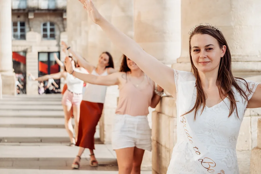 La mariée en robe de dentelle blanche, écharpe et diadème, bras levé devant ses amies alignées le long des colonnes de pierre, EVJF à Bordeaux