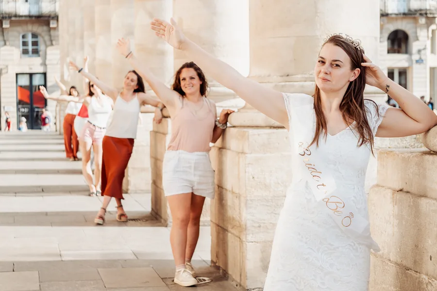 La mariée en robe de dentelle blanche, écharpe Bride to be et diadème, une main dans les cheveux, devant ses amies alignées bras tendus le long des colonnes, EVJF à Bordeaux