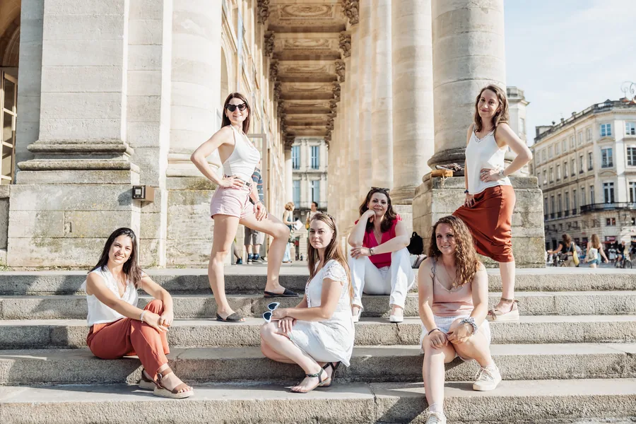 La mariée en robe de dentelle blanche et cinq amies sur des marches, au pied de hautes colonnes, quatre assises et deux debout, EVJF à Bordeaux
