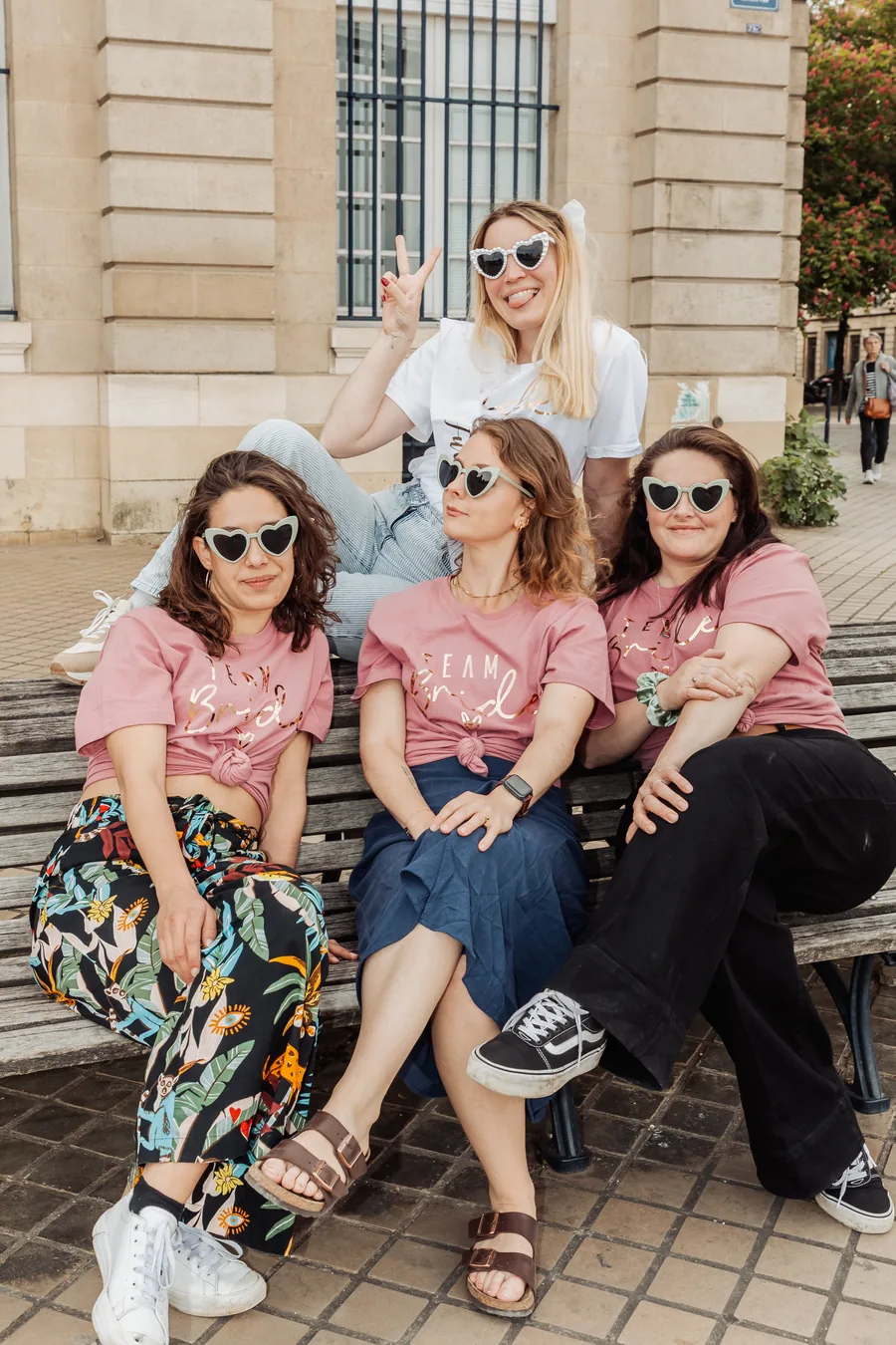 Trois amies en t-shirts roses et lunettes en cœur assises sur un banc, la mariée en t-shirt blanc perchée derrière, deux doigts levés, EVJF à Bordeaux
