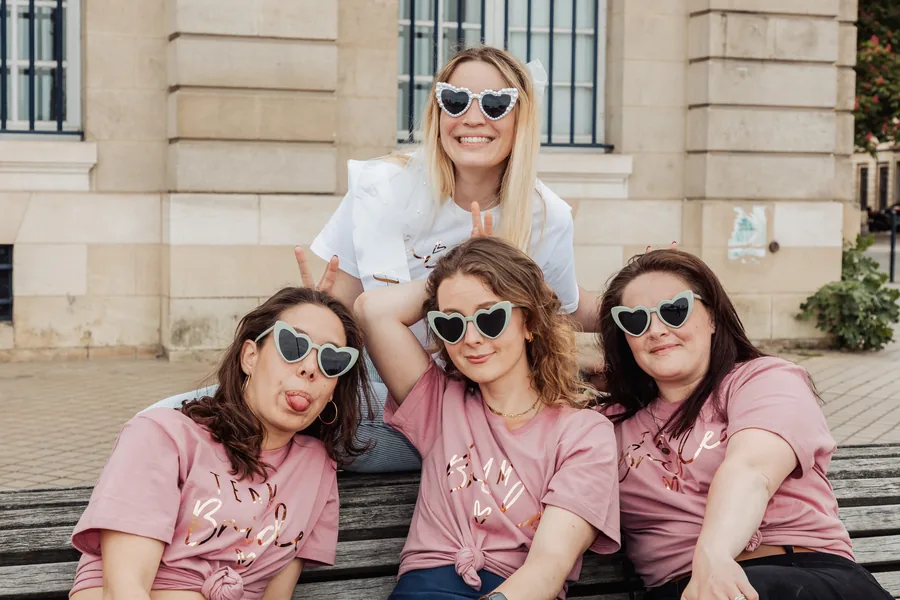 La mariée en t-shirt blanc fait des oreilles de lapin derrière ses trois amies en t-shirts roses, lunettes en cœur, sur un banc en bois, EVJF à Bordeaux