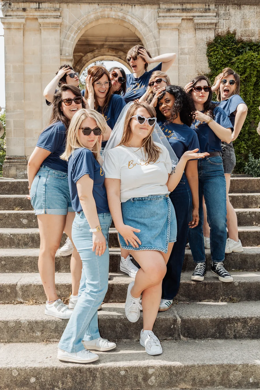 Dix amies en t-shirts bleu marine autour de la mariée en t-shirt blanc, voile et lunettes en cœur, sur des marches de pierre, EVJF à Bordeaux