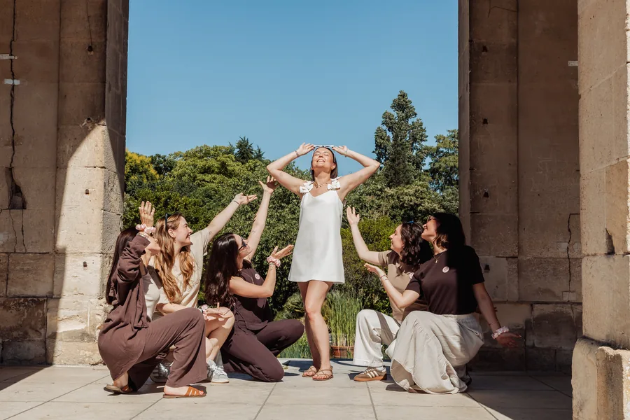 La mariée debout en robe blanche courte, cinq amies accroupies tendant les bras vers elle, entre deux piliers de pierre, sous un ciel bleu, EVJF à Bordeaux