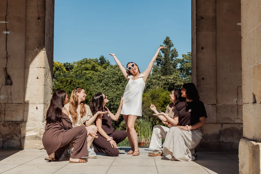 La mariée en robe blanche et lunettes en cœur, bras levés, entourée de cinq amies accroupies entre des piliers de pierre, EVJF à Bordeaux