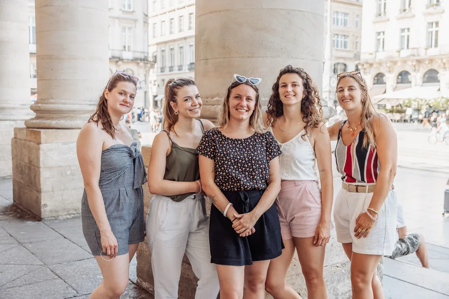Cinq amies debout sous les colonnes, tenues d'été, la place derrière elles, EVJF à Bordeaux