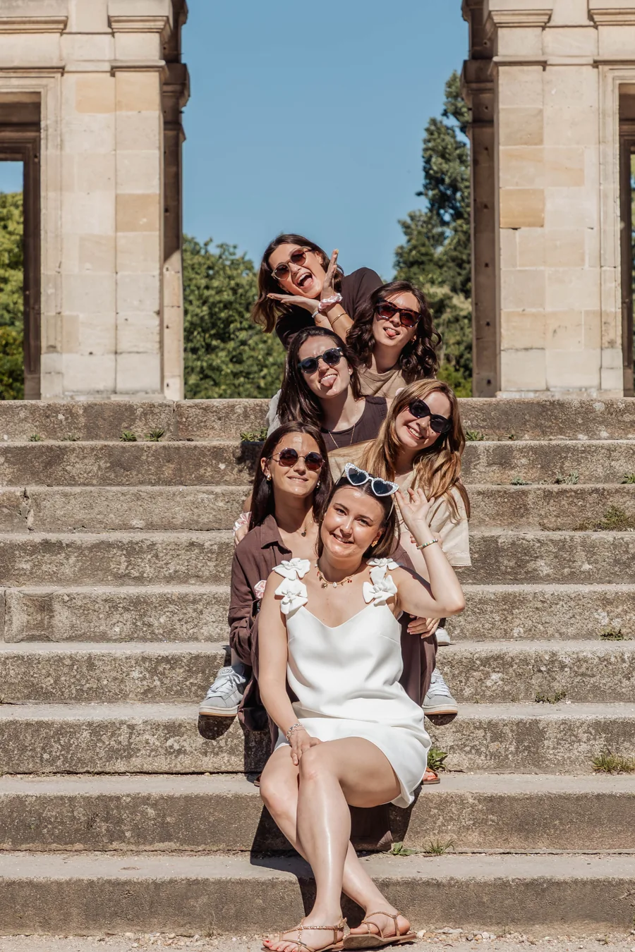 Six amies assises en file sur des marches de pierre, la mariée en robe blanche devant, grimaces et lunettes de soleil, EVJF à Bordeaux
