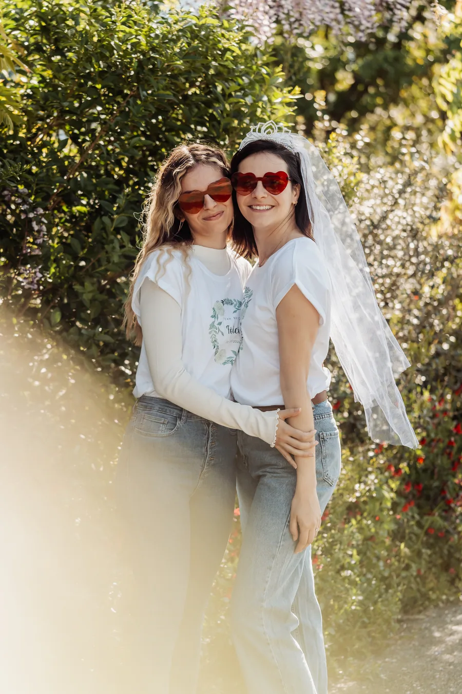 La mariée voilée et une amie enlacées, lunettes en cœur, dans un buisson fleuri à contre-jour, EVJF à Bordeaux