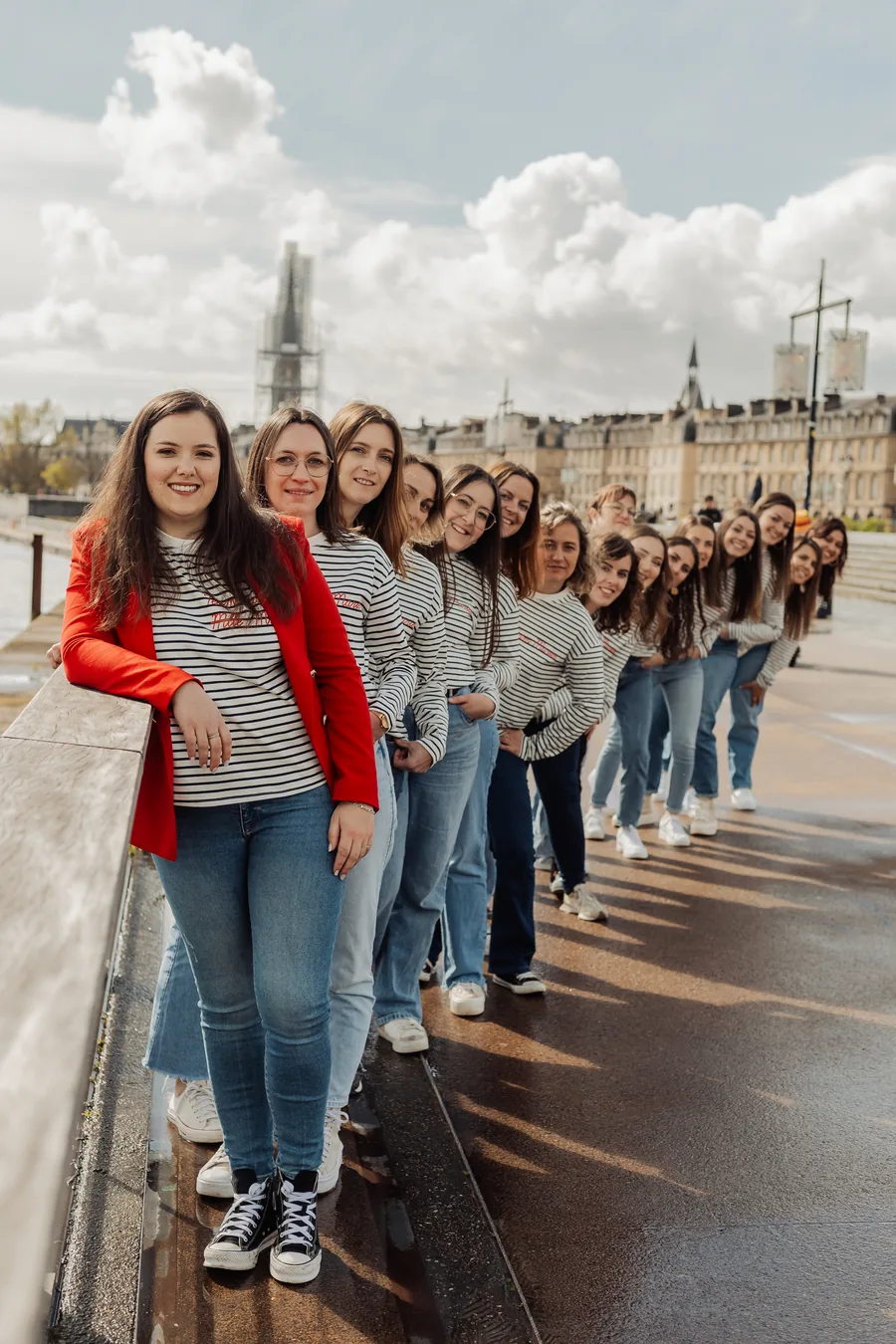 Une quinzaine d'amies en marinières alignées le long d'un parapet, têtes penchées vers l'objectif, veste rouge en tête, EVJF à Bordeaux