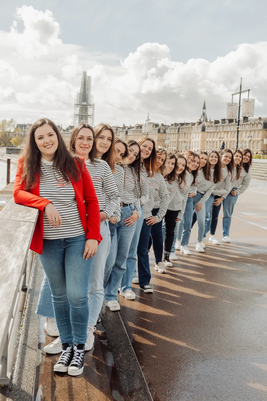 Le groupe en marinières et jean penché en file le long d'un parapet, sol mouillé et ciel nuageux, une veste rouge devant, EVJF à Bordeaux