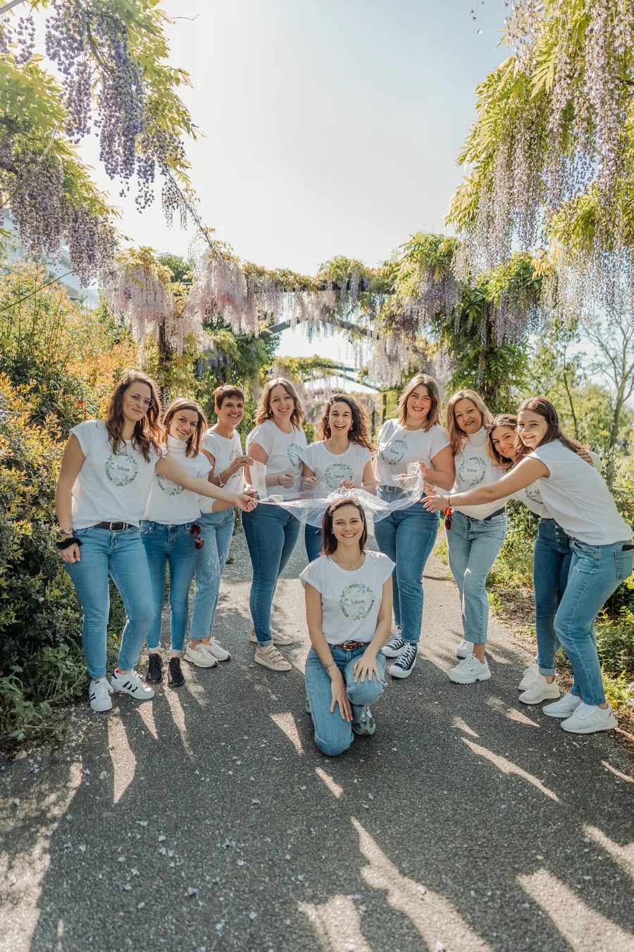 Neuf amies en t-shirts blancs et jean sous une pergola de glycine, tendant un voile derrière la mariée agenouillée, diadème sur la tête, EVJF à Bordeaux