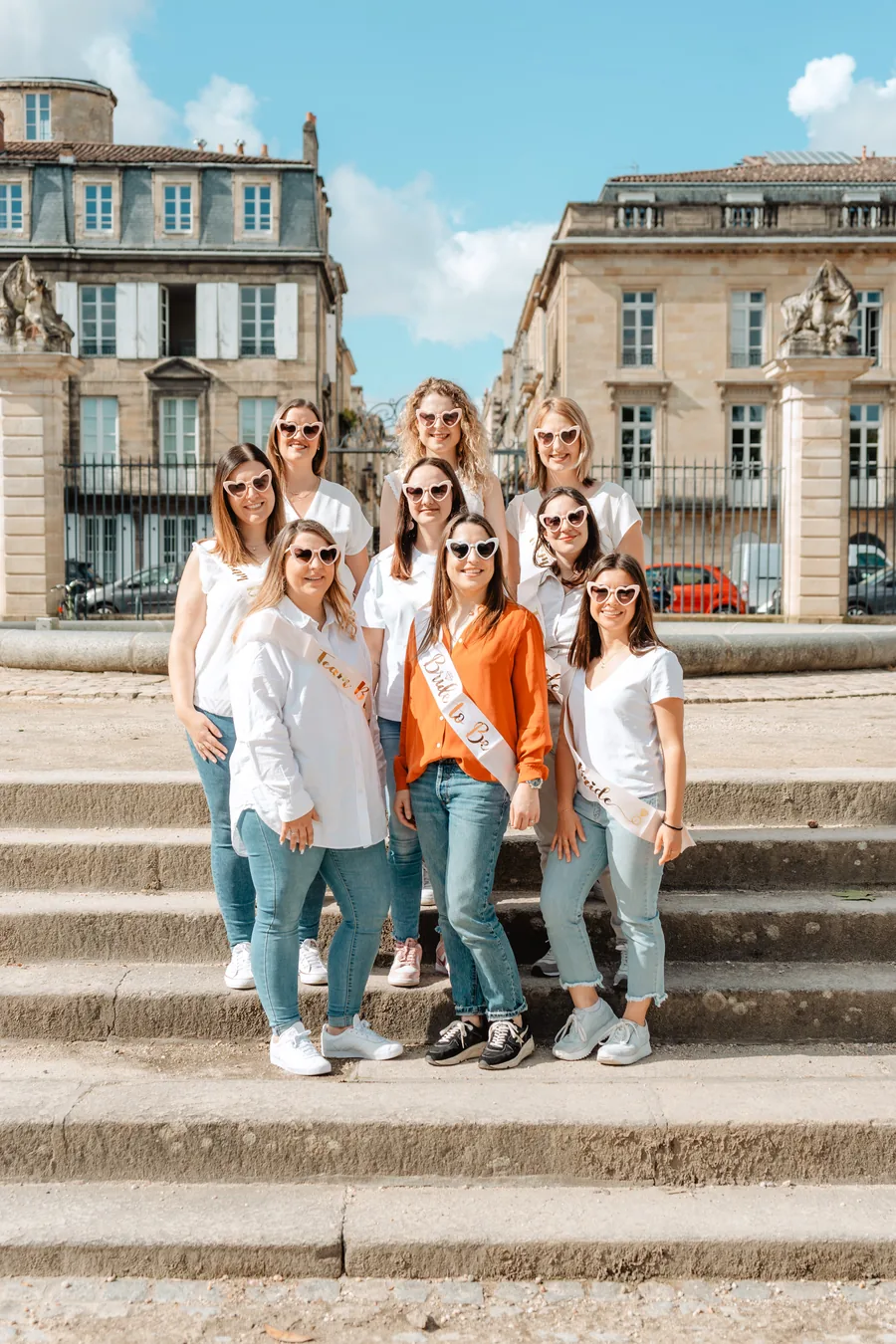 Neuf amies en lunettes en forme de cœur sur des marches de pierre, la mariée en chemise orange et écharpe au centre, EVJF à Bordeaux