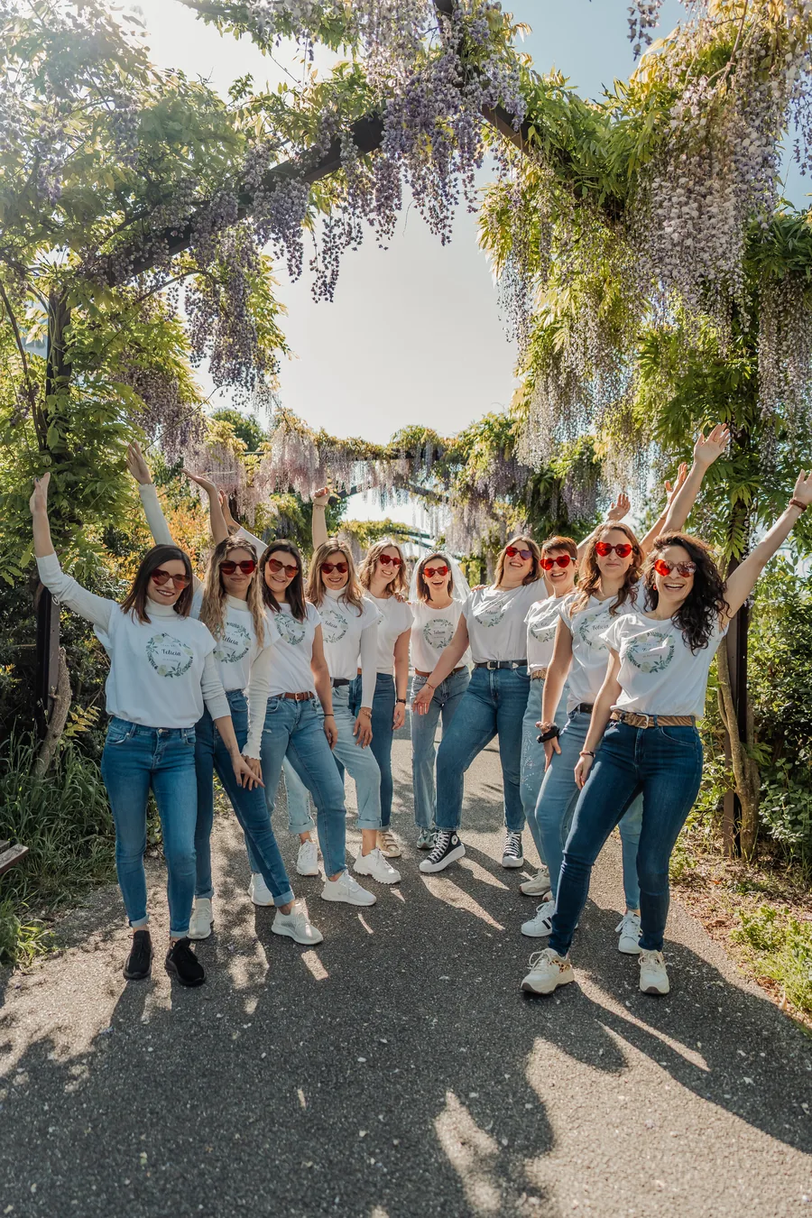 Dix amies sous une arche de glycine, la plupart bras levés, lunettes cœur rouges et t-shirts blancs assortis, la mariée voilée au centre, EVJF à Bordeaux