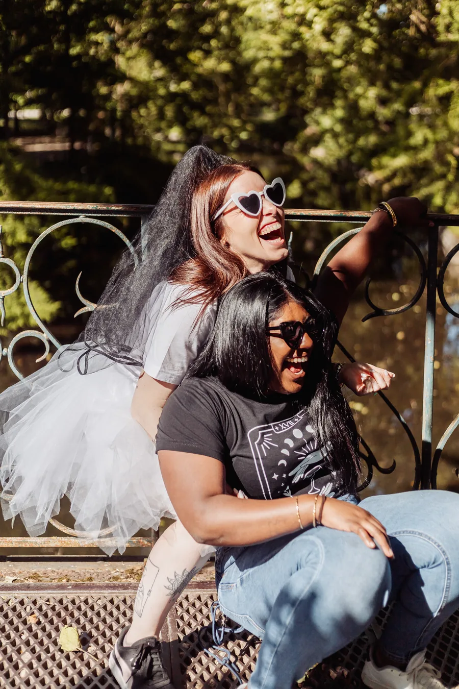 La mariée en voile noir, tutu blanc et lunettes en forme de cœur rit derrière une amie, contre une rambarde en fer forgé, EVJF à Bordeaux