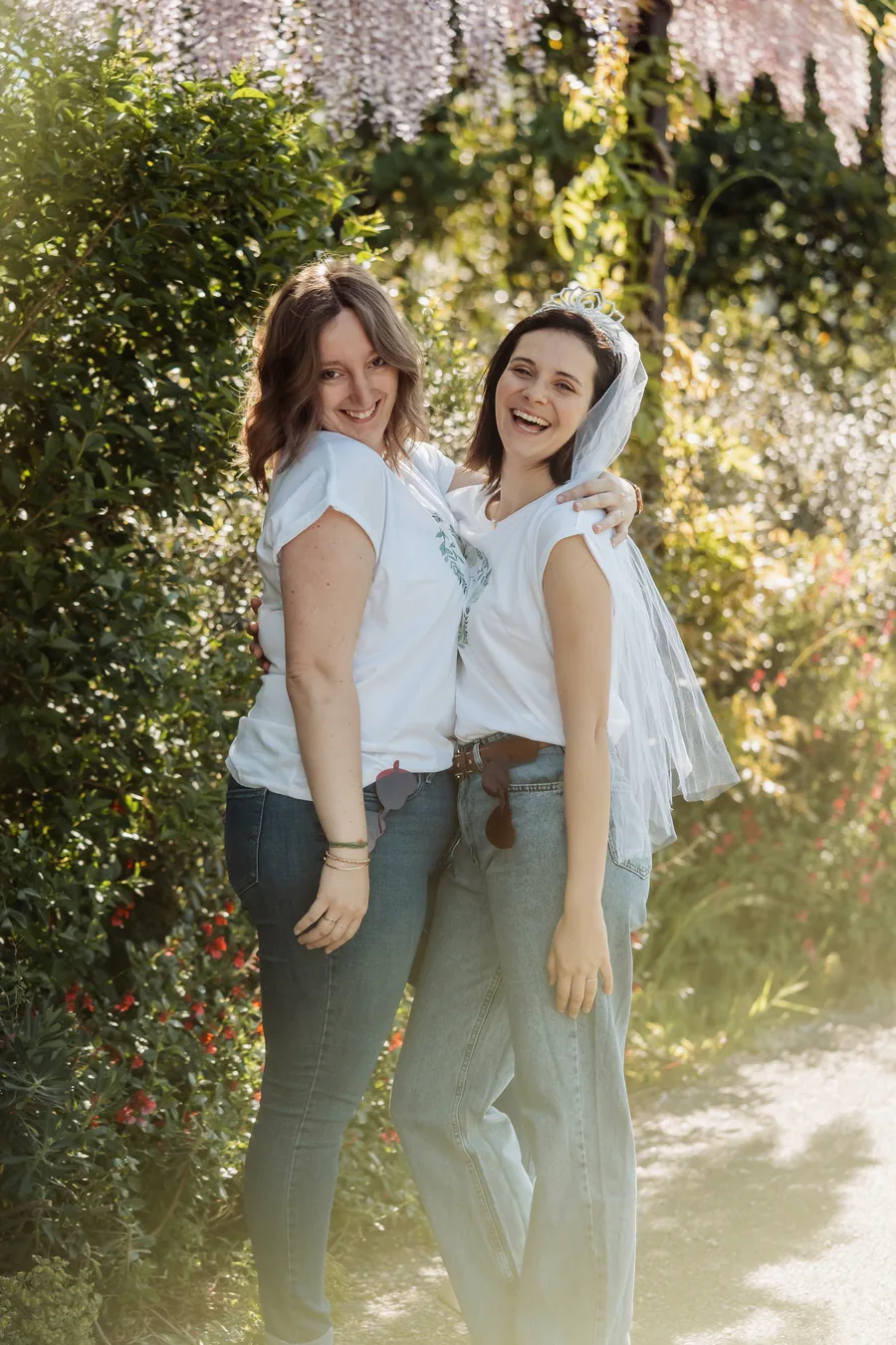 La mariée voilée et une amie enlacées, lunettes cœur accrochées à la ceinture, devant des buissons fleuris à contre-jour, EVJF à Bordeaux