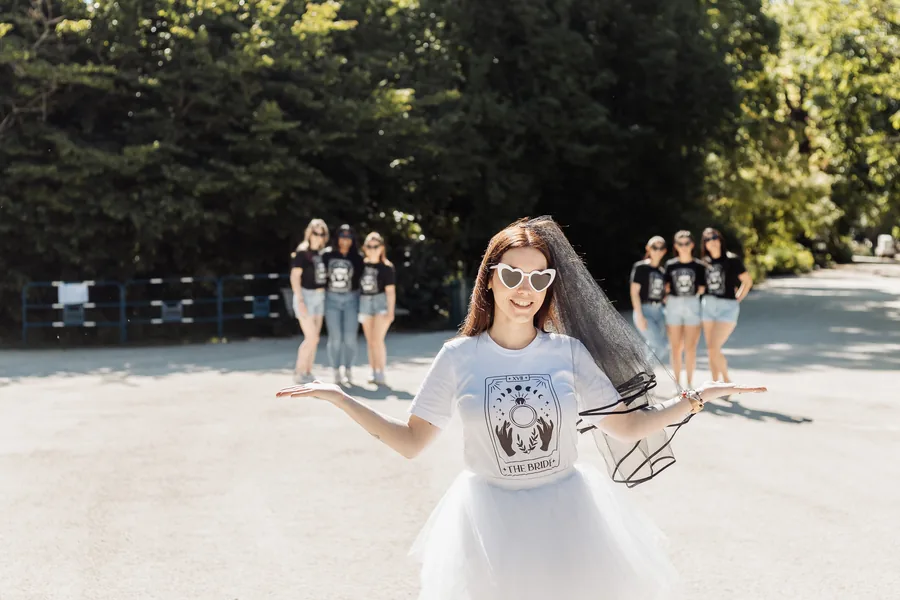 La mariée en voile noir, tutu blanc et lunettes cœur, bras ouverts, six amies en t-shirts noirs floues à l'arrière-plan, EVJF à Bordeaux