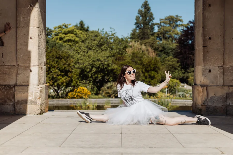La mariée en tutu blanc et lunettes en forme de cœur, en grand écart sur les dalles entre deux piliers de pierre, jardin en fond, EVJF à Bordeaux