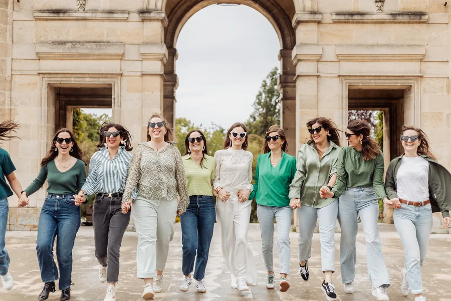 Le groupe court main dans la main vers l'objectif, lunettes en forme de cœur, la mariée en blanc au centre, devant une arche de pierre, EVJF à Bordeaux