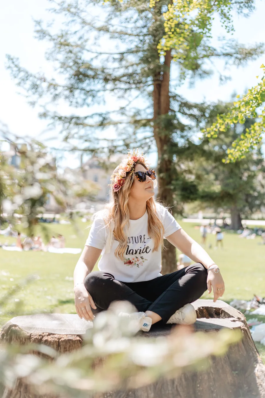 La mariée en couronne de fleurs et lunettes de soleil, assise en tailleur sur une souche dans un parc, grand conifère en fond, EVJF à Bordeaux