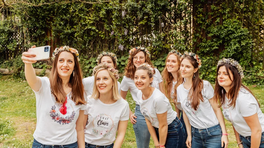 Huit amies en couronnes de fleurs et t-shirts blancs assortis prennent un selfie dans un jardin, devant un mur de plantes grimpantes, EVJF à Bordeaux