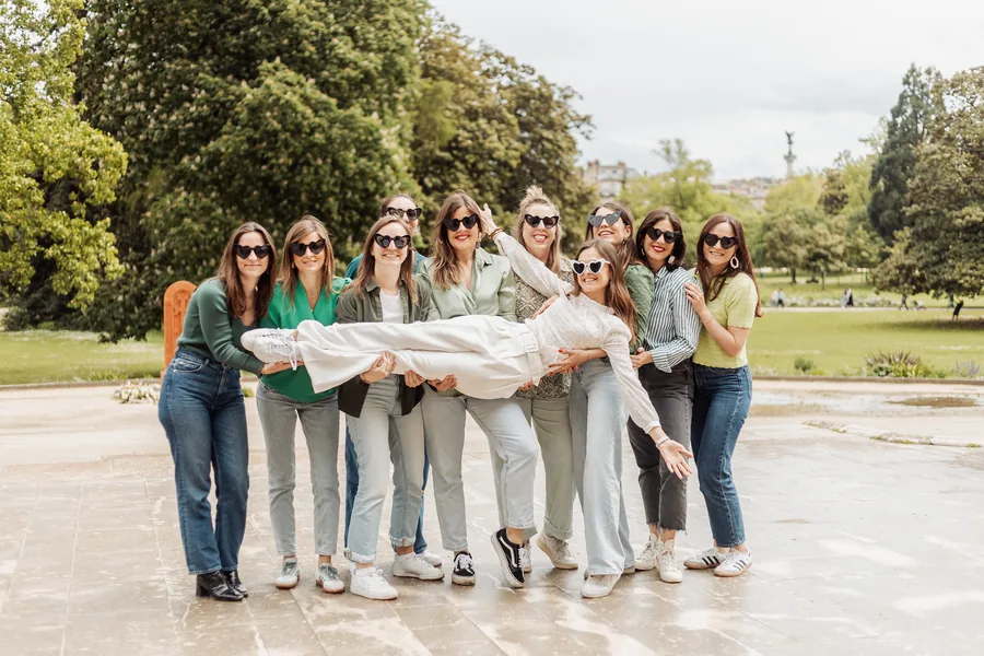 Neuf amies portent à l'horizontale la mariée vêtue de blanc, lunettes en forme de cœur, sur l'esplanade d'un parc arboré, EVJF à Bordeaux