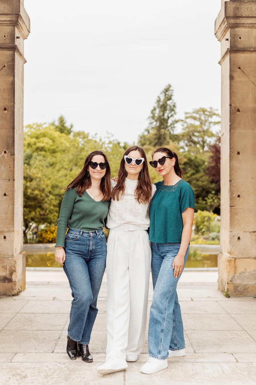 La mariée en blanc enlacée par deux amies, lunettes en forme de cœur, debout entre deux piliers de pierre, jardin en fond, EVJF à Bordeaux