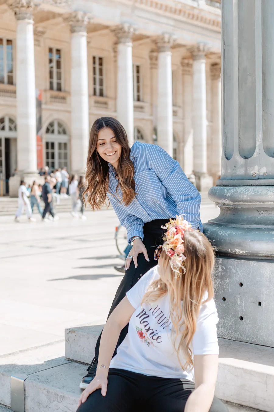 Une amie en chemise rayée se penche vers la mariée assise sur des marches, couronne de fleurs, devant une façade à colonnes, EVJF à Bordeaux
