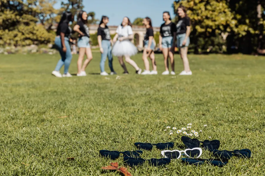 Lunettes en forme de cœur posées dans l'herbe près de pâquerettes, six amies et la mariée en tutu blanc floues à l'arrière-plan, EVJF à Bordeaux