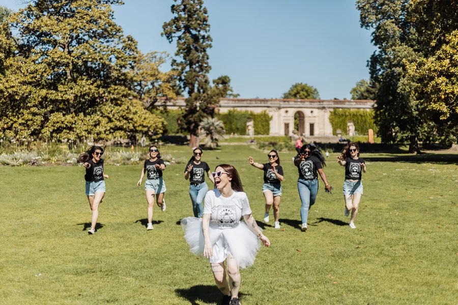 La mariée en tutu blanc et lunettes en cœur court en riant sur la pelouse, six amies en t-shirts noirs à ses trousses, EVJF à Bordeaux