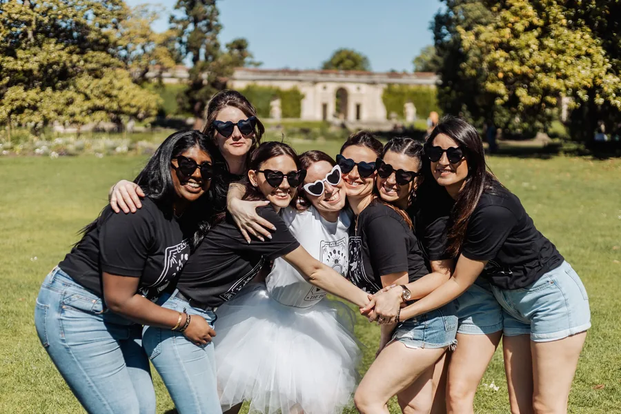 Six amies serrées autour de la mariée en tutu blanc, mains réunies devant elles, lunettes en forme de cœur, sur une pelouse, EVJF à Bordeaux