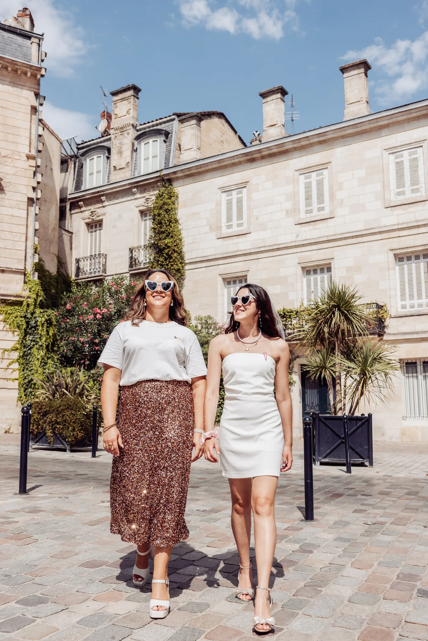 Deux copines qui marchent côte à côte devant une façade bordelaise, robe blanche et jupe à paillettes, EVJF à Bordeaux
