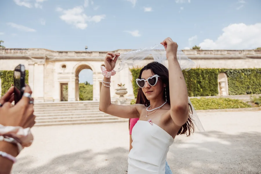 La mariée en bustier blanc et lunettes cœur à perles soulève son voile, photographiée au téléphone devant une arche de pierre, EVJF à Bordeaux