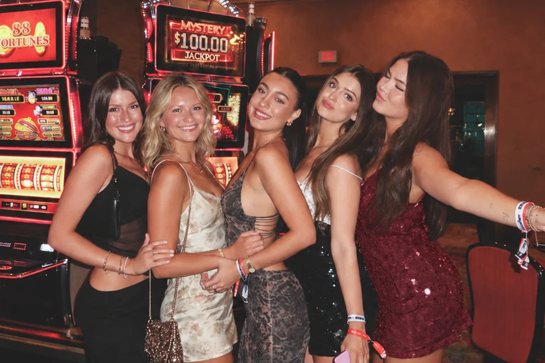Cinq copines en robes de soirée posent enlacées devant des machines à sous