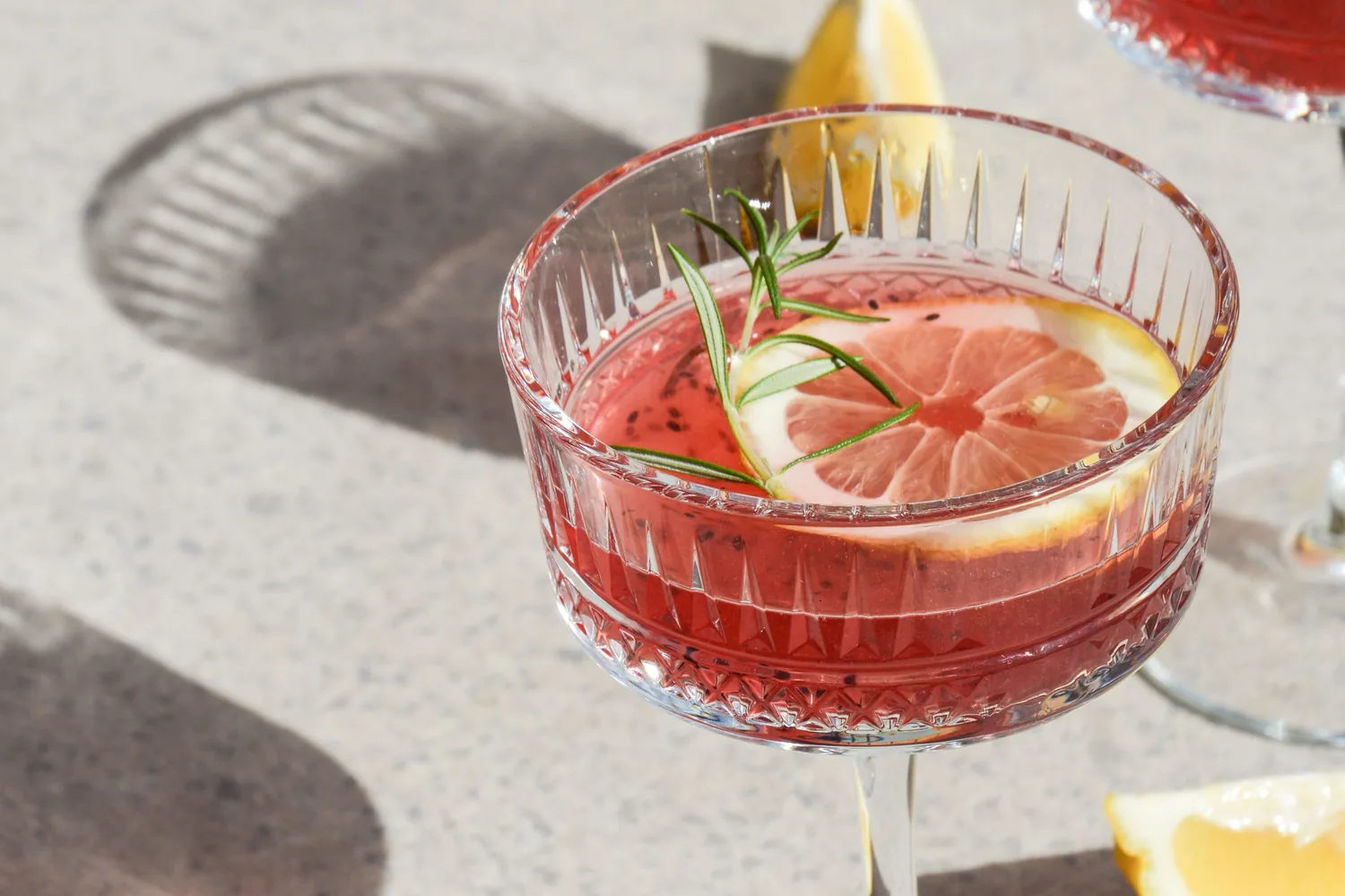 Une coupe de cocktail rose, rondelle d'agrume et brin de romarin, au soleil sur une table claire