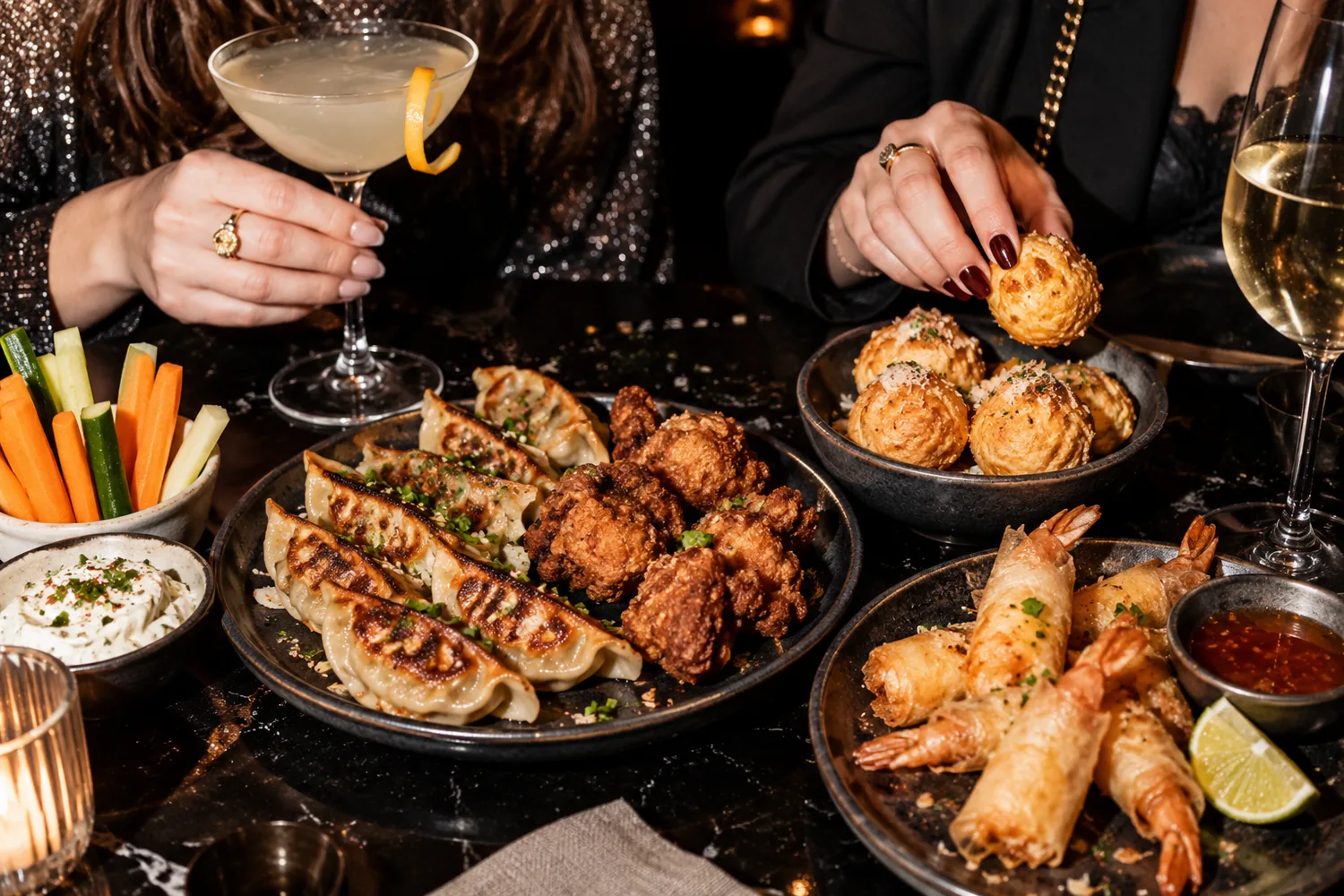 Un cocktail dans une coupe, un verre de vin blanc et des plateaux de bouchées : raviolis grillés, poulet frit, crevettes en feuille de brick, crudités