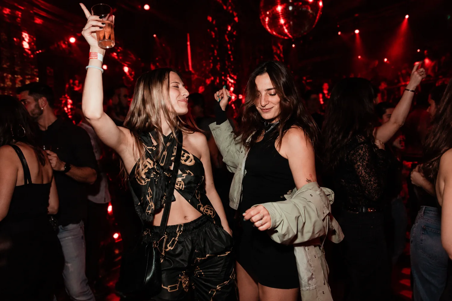 Deux amies dansent sous une lumière rouge, un verre levé, une boule à facettes au plafond