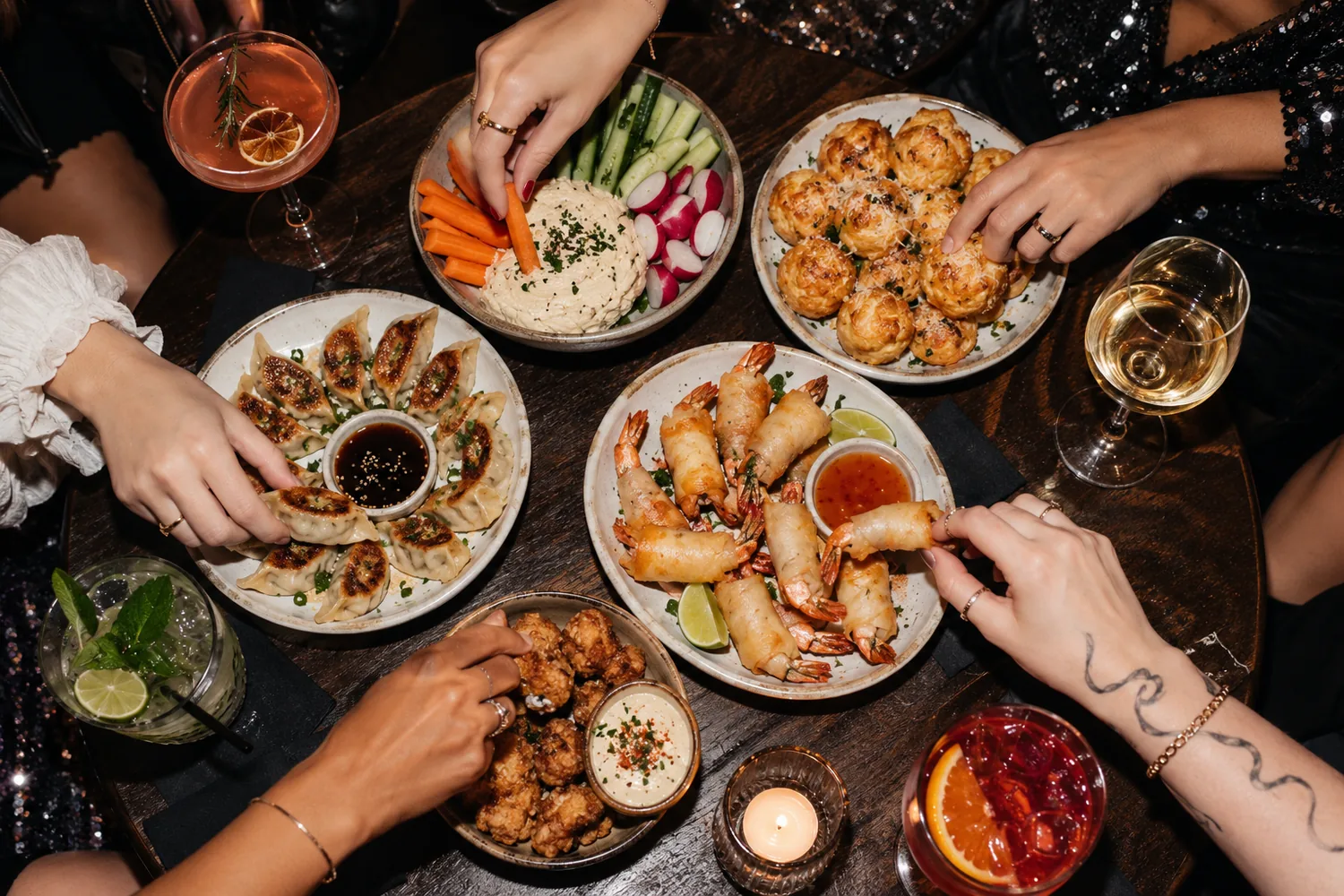 Vue du dessus d'une table ronde : des mains piochent dans les raviolis, les crevettes, les bouchées au fromage et les crudités, cocktails autour