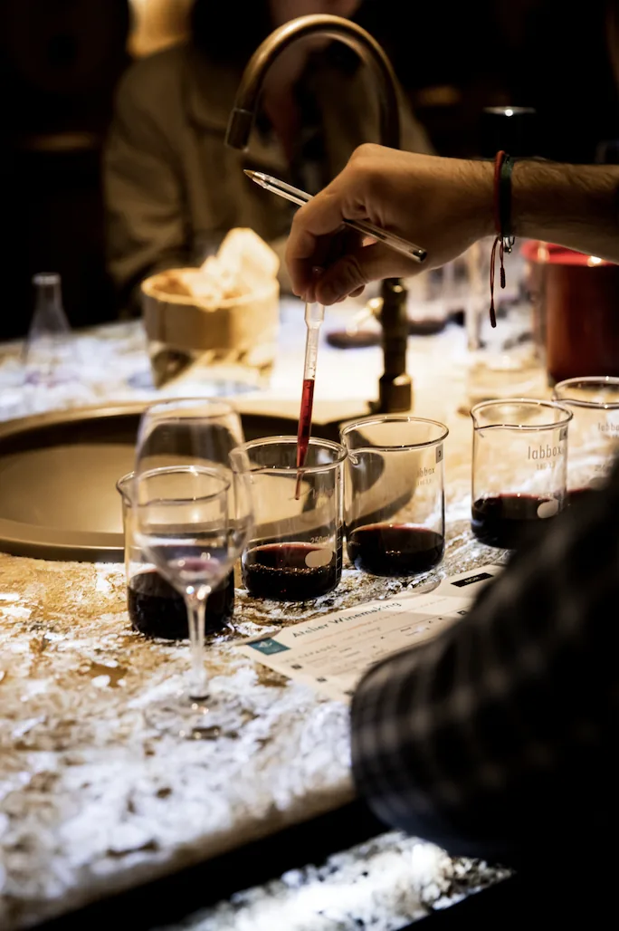 Une main dose le vin à la pipette dans des béchers gradués, verres et fiche de dégustation posés autour, EVJF à Bordeaux