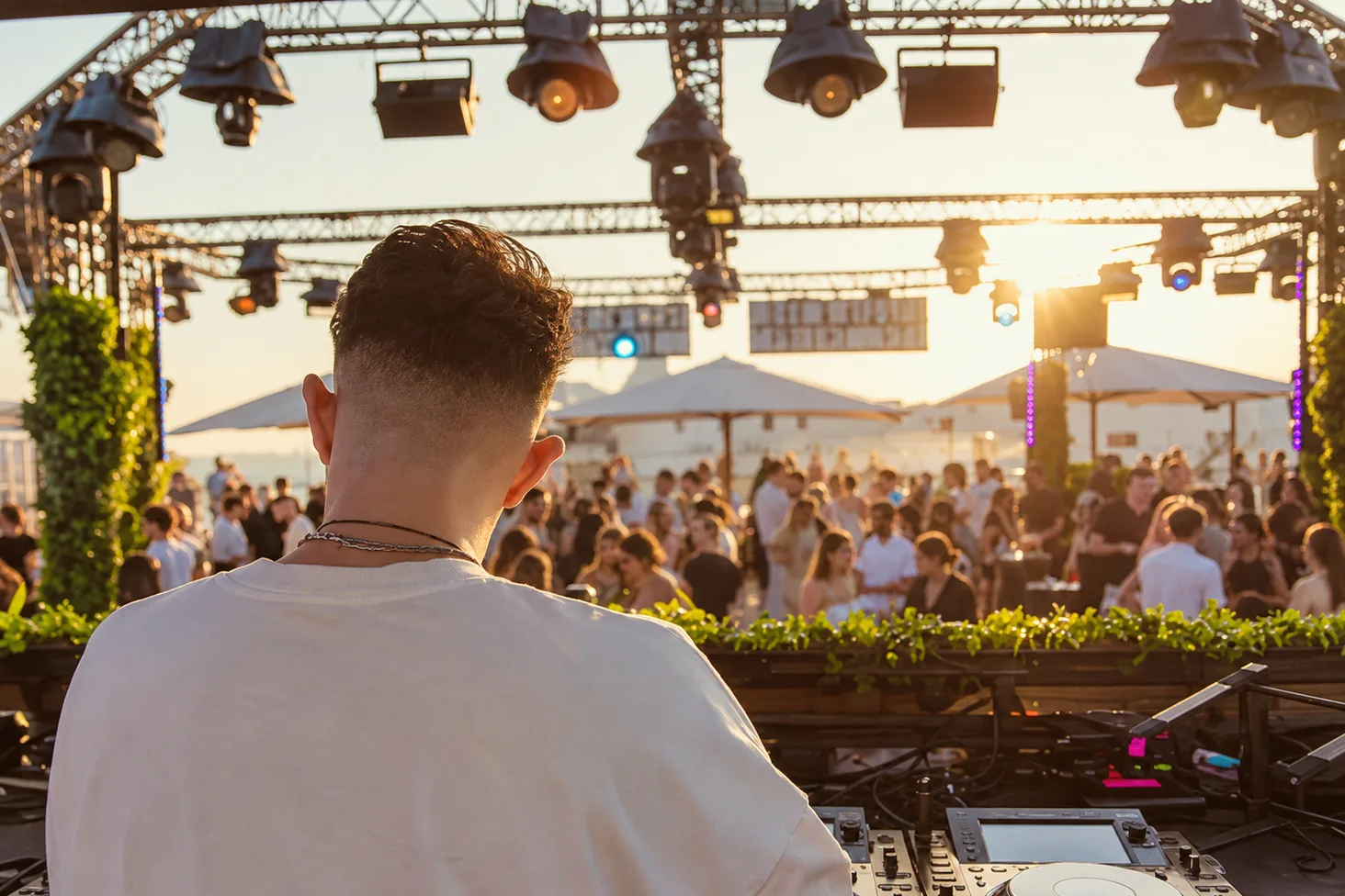 Un DJ vu de dos aux platines, face à la foule d'une terrasse, au coucher du soleil