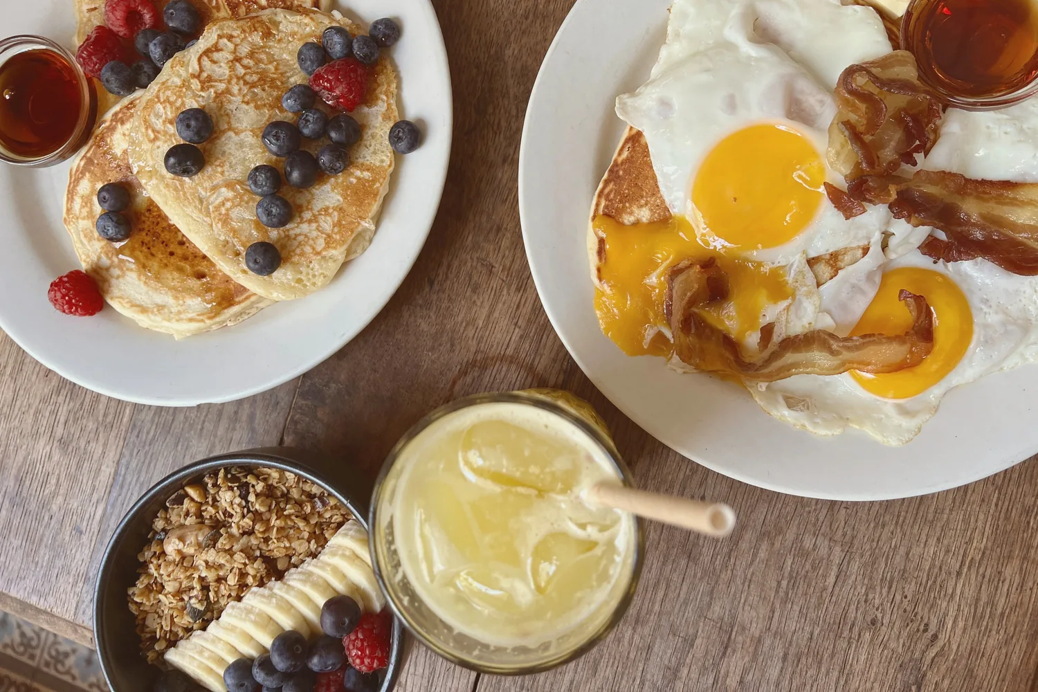 Brunch vu du dessus sur une table en bois : pancakes aux myrtilles et framboises, œufs au plat et bacon, granola aux fruits et jus pressé