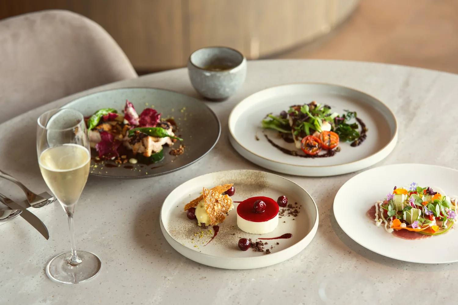 Quatre assiettes dressées et une flûte de champagne sur une table ronde : entrée fleurie, poisson, volaille et dessert aux fruits rouges
