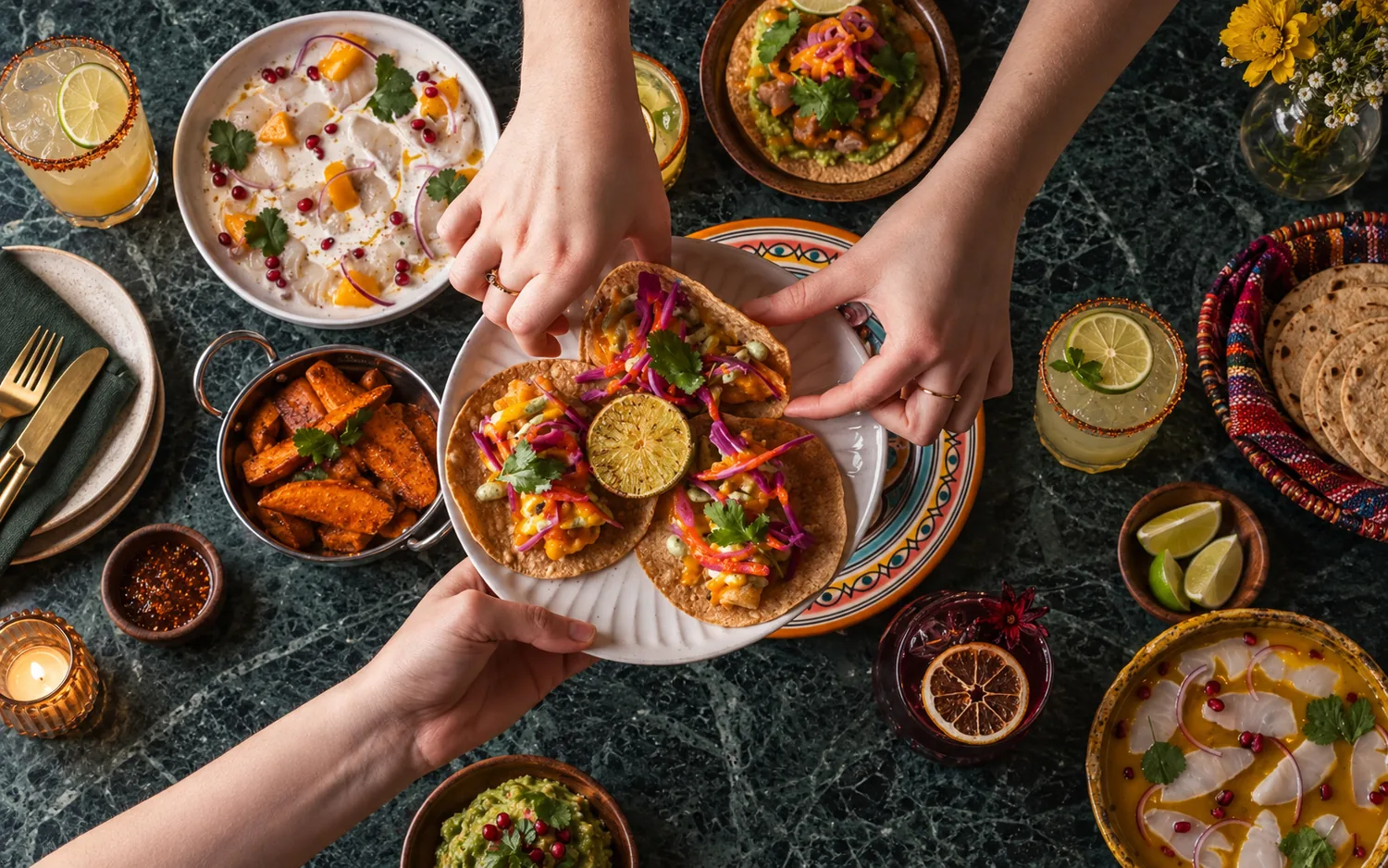 Vue du dessus d'une table à partager sur un marbre vert : tacos passés de main en main, ceviche, patates douces, guacamole et cocktails au citron vert