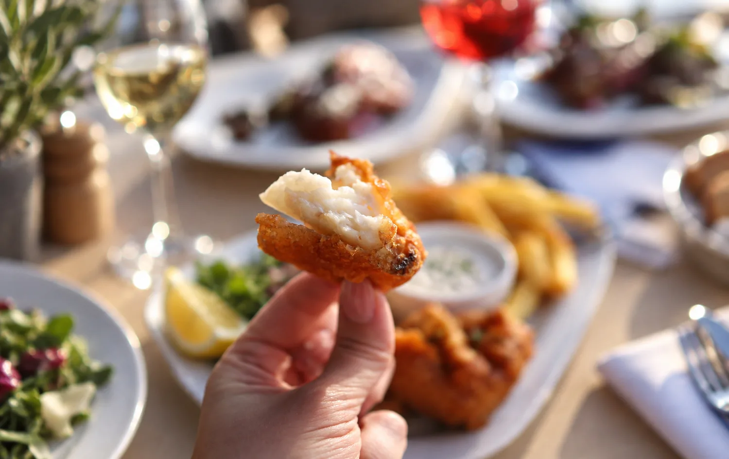 Une bouchée de poisson pané tenue du bout des doigts devant un fish and chips, sa sauce blanche et un quartier de citron, verres de vin en arrière-plan
