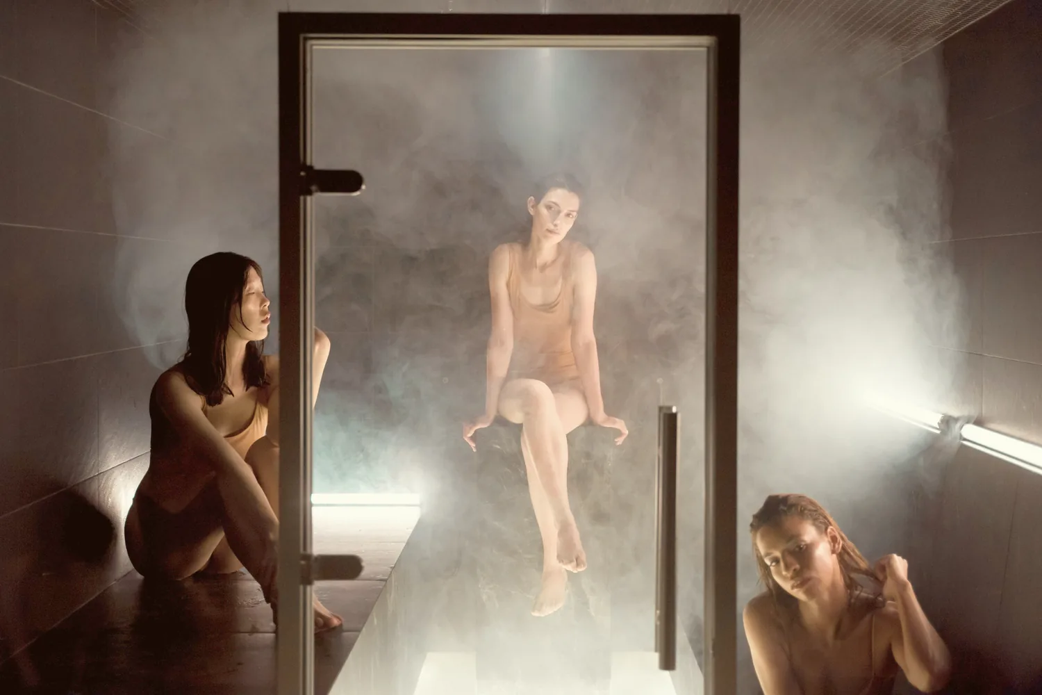 Trois femmes en maillot dans la vapeur d'un hammam, vues à travers la porte vitrée